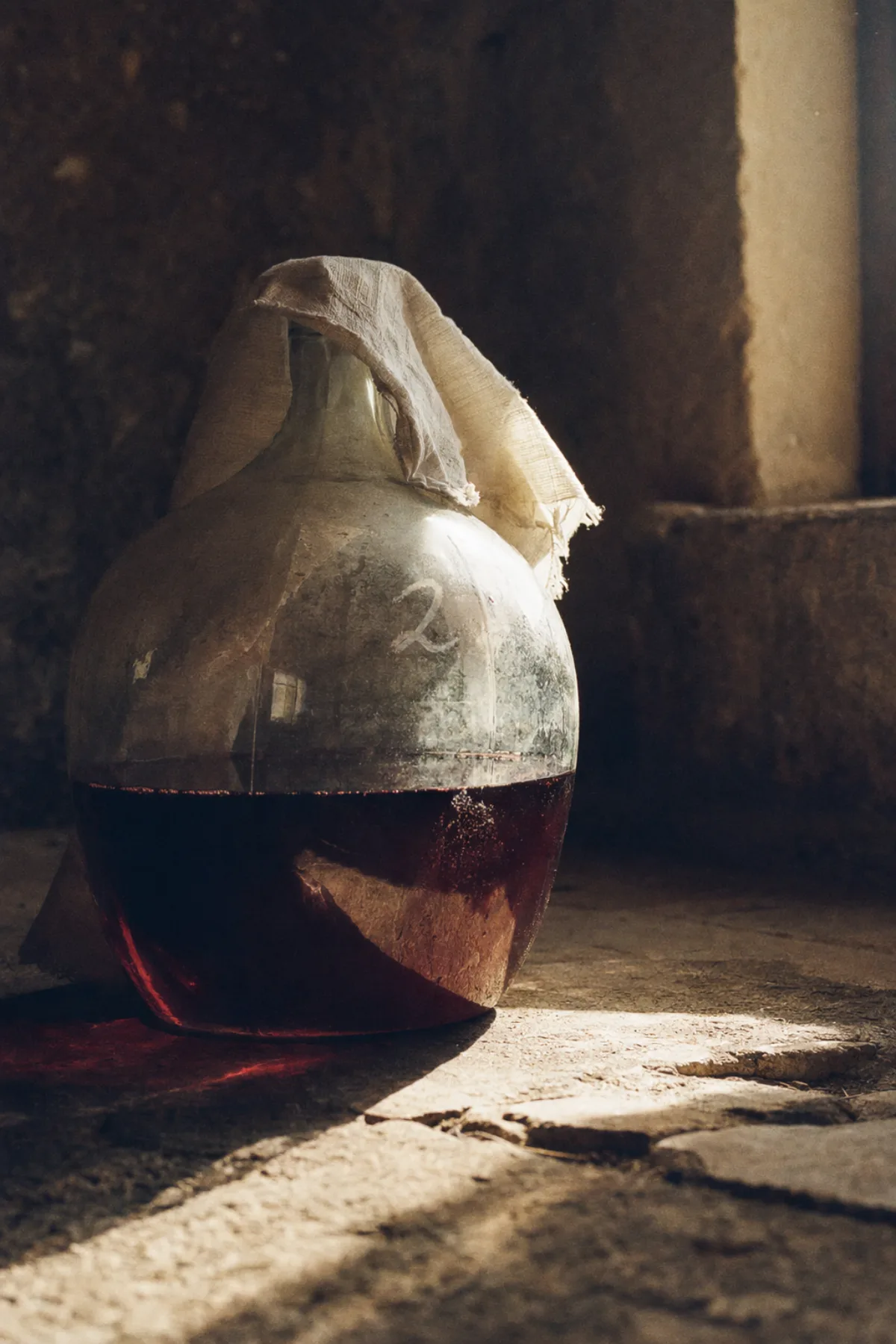 A glass demijohn half full of ruby-dark wine on a stone floor