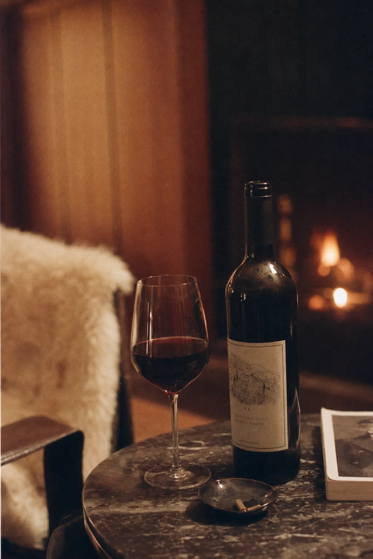 A half-full glass of dark cabernet on a side table, fire out of focus behind